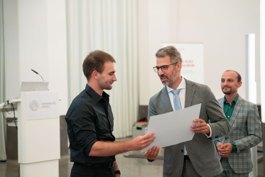 zur Vergrößerungsansicht des Bildes: Das Bild zeigt drei junge Männer, die zusammen auf einer Bühne stehen. Die mittlere Person überreicht der links stehenden Person mit beiden Händen einen Scheck. 