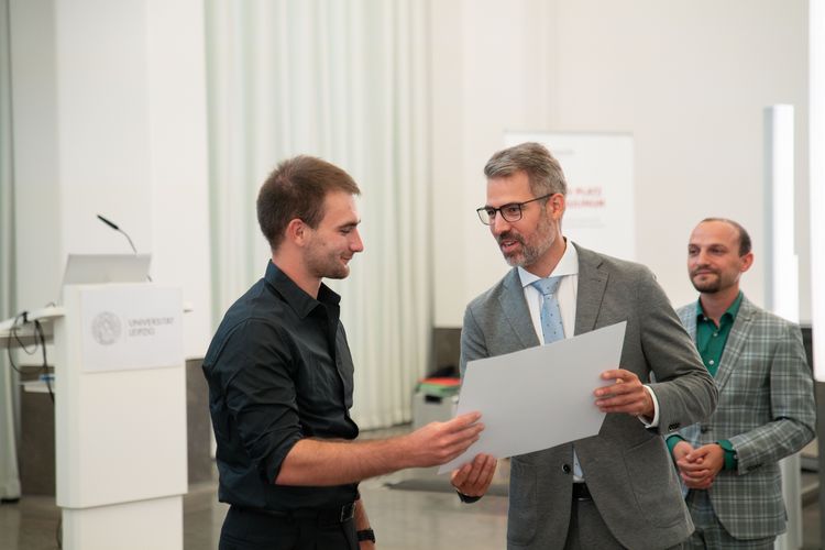 Das Bild zeigt drei junge Männer, die zusammen auf einer Bühne stehen. Die mittlere Person überreicht der links stehenden Person mit beiden Händen einen Scheck. 