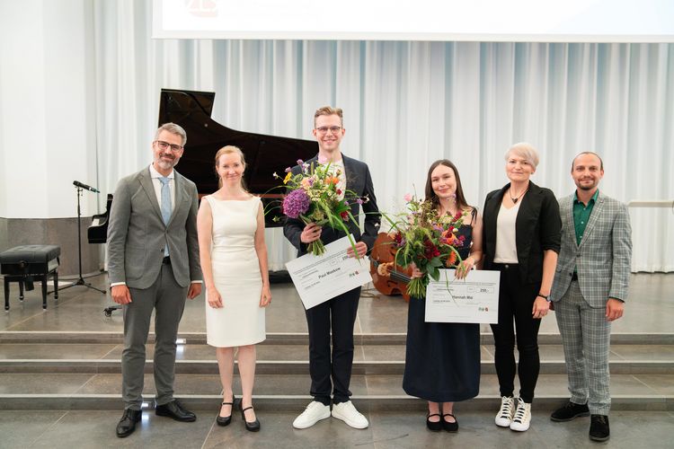 Das Foto zeigt sechs Menschen, die nebeneinander auf einer Bühne stehen und in die Kamera schauen. Ganz links und ganz rechts stehen zwei Herren in grauem Anzug, neben ihnen jeweils zwei blonde Frauen. In der Mitte stehen ein junger Mann und eine Frau, die jeweils einen Blumenstrauß sowie einen großen Scheck in den Händen halten. Im Hintergrund steht linker Hand ein Flügel. 