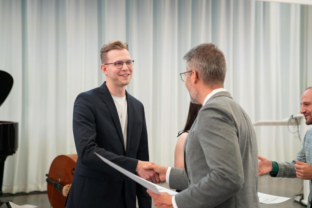 zur Vergrößerungsansicht des Bildes: Das Foto zeigt einen jungen Mann, der einem Mann von hinten die Hand schüttelt. Die Person, die mit dem Rücken zur Kamera steht, überreicht mit der linken Hand eine Urkunde. 