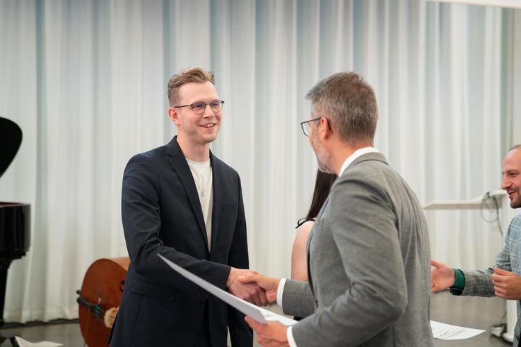 Das Foto zeigt einen jungen Mann, der einem Mann von hinten die Hand schüttelt. Die Person, die mit dem Rücken zur Kamera steht, überreicht mit der linken Hand eine Urkunde. 