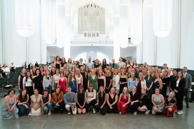 Das Foto zeigt ca. 100 junge Menschen, die in einem Pulk zusammenstehen, mit dem Blick in die Kamera gerichtet. Hinter ihnen deutet sich eine große, lichtdurchflutete Aula und eine Orgel an. 