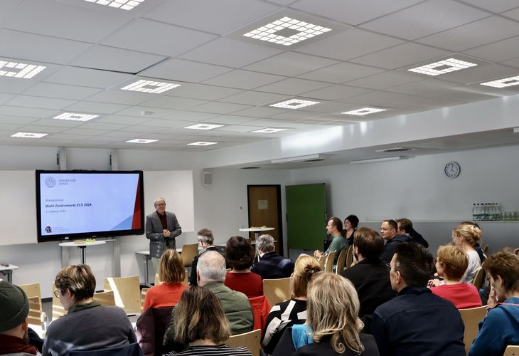 Das Foto zeigt eine redende Person, die im vorderen Bereich eines Raumes vor einem laufenden Smartboard steht und dem Publikum gegenüber eine Rede hält. 