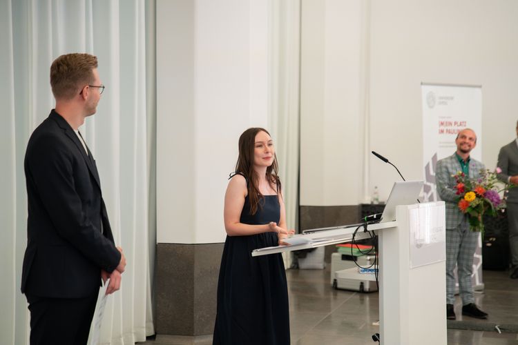 Das Foto zeigt drei Menschen, die etwa ein bis zwei Meter voneinander weg auf einer Bühne stehen. Mittig steht eine junge Frau, die vor einem Pult an einem Mikrofon eine Rede hält. Links neben ihr steht ein junger Mann - mit Blick auf sie gerichtet - der seine Hände vor dem Oberkörper zusammenhält. Rechts befindet sich ein Mann, der vor einem Roll-up steht und ebenfalls den Blick auf die Rednerin gerichtet hat. 